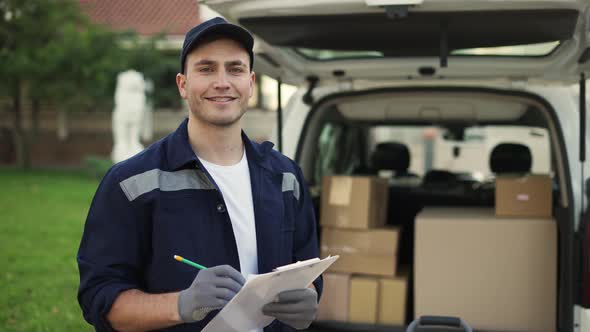 Happy Smiling Delivery Service Worker in Uniform Cap and Gloves Makes Notes on Documents and