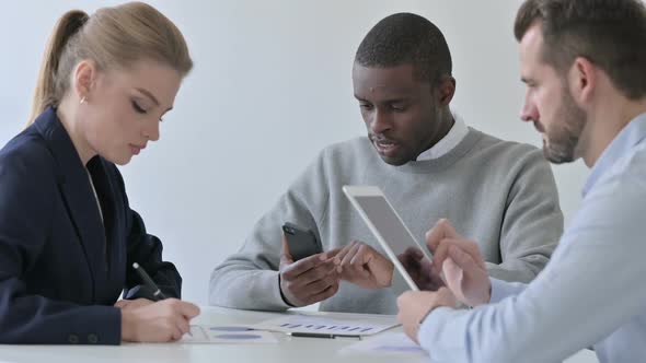 Businesswoman Writing on Paper While Male Colleagues Research on Smartphone and Tablet