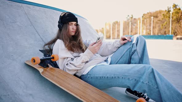 Casual Dressed Teenage Girl Lies on a Springboard in a Skatepark Listening to Music