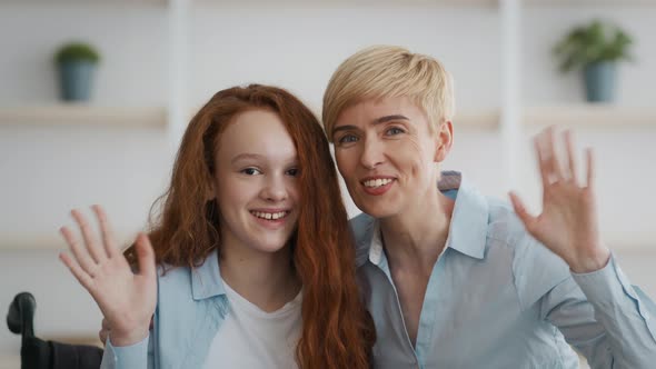 Little Handicapped Redhead Girl Sitting in Wheelchair Embracing with Her Mother and Waving Hand to