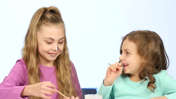 Two Little Girls Paint Their Face with Different Colors. Close Up. White Background alt