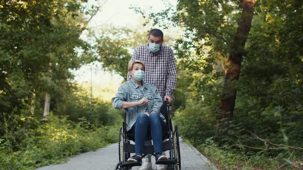Disabled Person Woman in Wheelchair with Her Man Stand on the Path in Park Wearing Masks to Protect