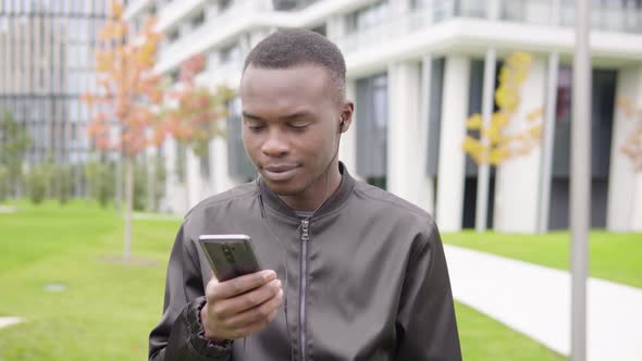 A Young Black Man Listens To Music with Earphones on a Smartphone - Office Buildings