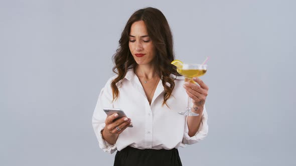 Lady in White Shirt and Black Skirt is Typing on Her Smartphone and Holding Cocktail While Posing on