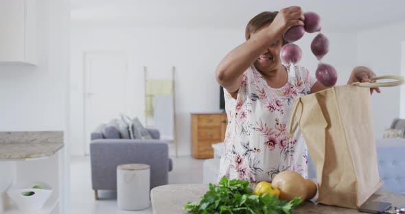 Happy african american senior woman in kitchen unpacking food from shopping bag, smiling