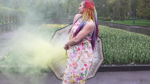 Girl with African Braids and Makeup in Spring Dress is Dancing in the Rain with Umbrella in Yellow