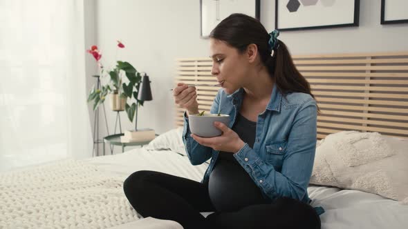 Tracking video of caucasian woman in advanced pregnancy sitting on bed, eating salad