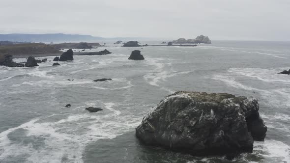 Stormy day aerial of a rocky ocean shore, tracking over a large rock in the ocean, waves crash again