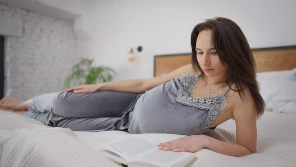 Young Intelligent Pregnant Woman Reading Book Lying on Bed at Home