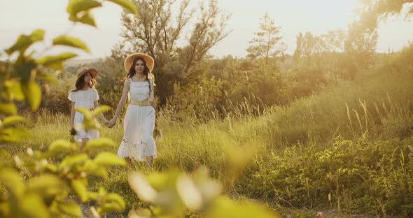 Two Girl Teenagers in White Dresses and Straw Hats with Flowers Bouquet Walking on Nature at Sunny