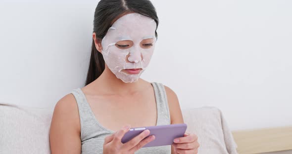 Woman apply facial paper mask on face and use of mobile phone at home