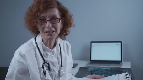 Senior Woman Doctor Speaks Looking Into Camera Holding Clipboard in Hands Sitting in Medical Office