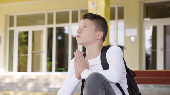 A Caucasian Teenage Boy Prays with His Hands Clasped Together As He Sits in Front of School