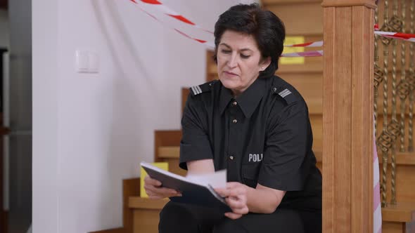 Concentrated Caucasian Policewoman Examining Photo Evidence Sitting on Stairs at Crime Scene