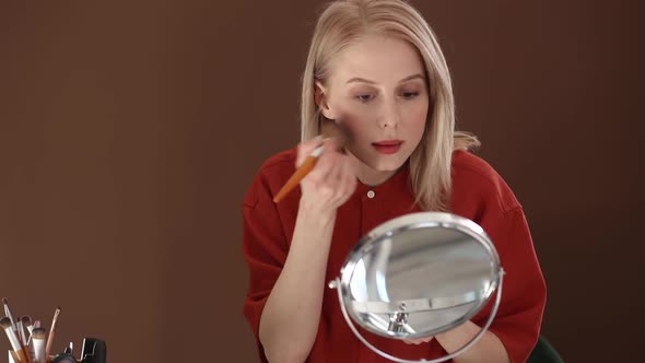 Beautiful blond hair woman in red shirt applying makeup in opposite of mirror