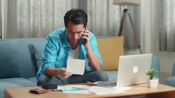 Asian Man With A Laptop Answering The Call And Having Headache While Looking At The Bill In Hand