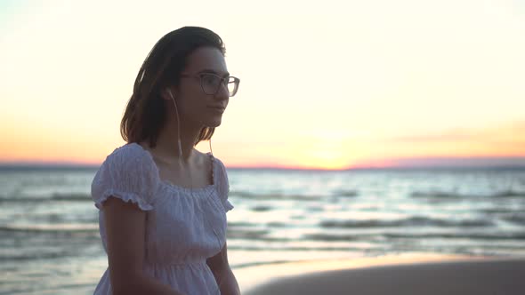 A Young Woman Sits on a Stone on the Beach By the Sea with Headphones in Her Ears. A Girl in a White