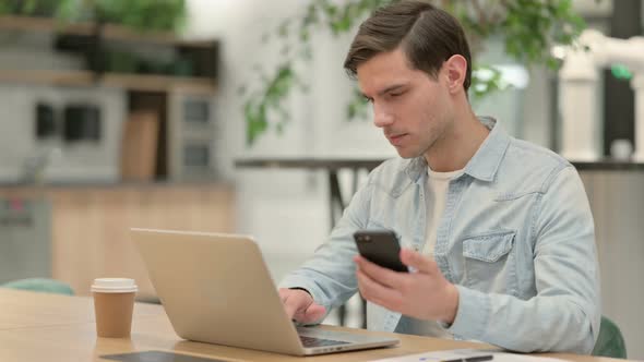 Creative Young Man with Laptop Using Smartphone at Work