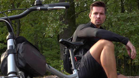 A Young Handsome Cyclist Sits on a Log Next To His Bike in a Forest and Looks Seriously