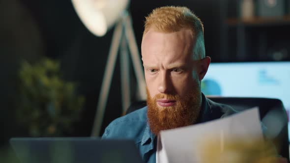 Bearded Hipster Guy Analyzing Data on Laptop and Documents in Office.