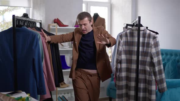 Satisfied Cheerful Gay Man Dancing in Slow Motion in Shop Admiring Clothing in Rack Hangers