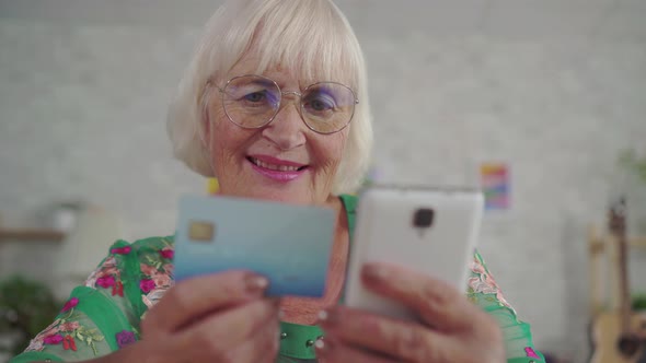 Elderly Woman with Gray Hair Holds a Bank Card and Uses a Smartphone