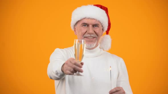 Glad Senior Man in Santa Claus Hat With Bengal Light Showing Champagne Glass