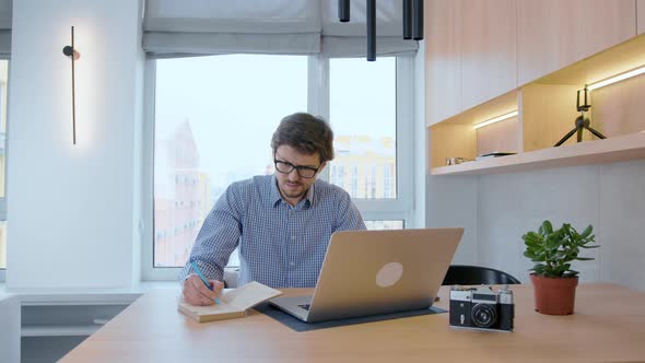 Man in glasses works at a laptop against the background of a window. Freelancer working remotely.
