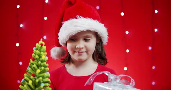 Cute Girl in a Santa Hat Holds a Gift on a Red Christmas Background