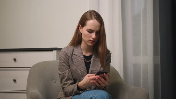 Upset Young Woman Sitting on Couch Holding Smartphone in Hands Received Message with Bad News