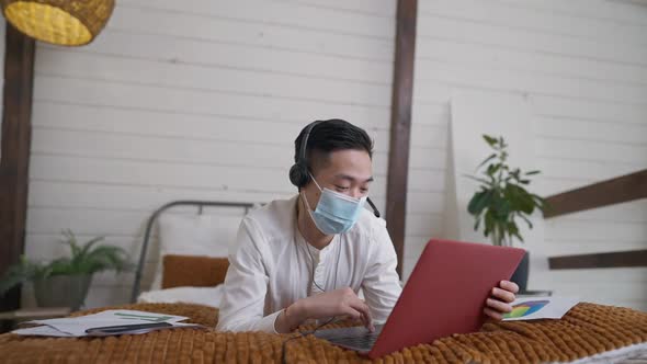 Positive Happy Young Chinese Man in Earphones and Covid Face Mask Messaging Online Surfing Internet