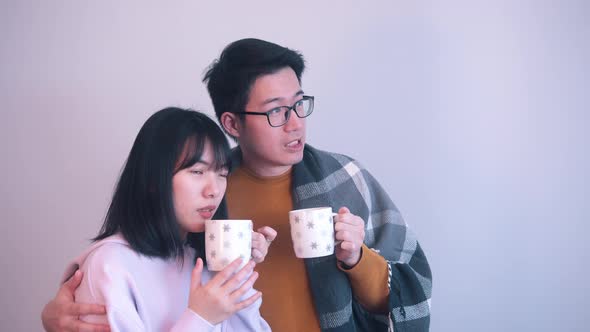Young Asian Couple Hugging Under the Blanket and Drinking Hot Beverage Indoors on Stormy Day