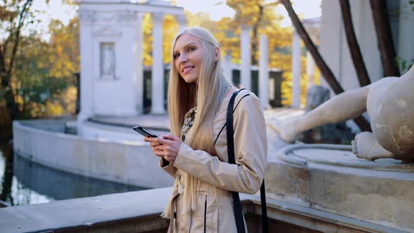 Cheerful Blonde Woman in Beige Coat in Autumn Park Looking at Her Smartphone