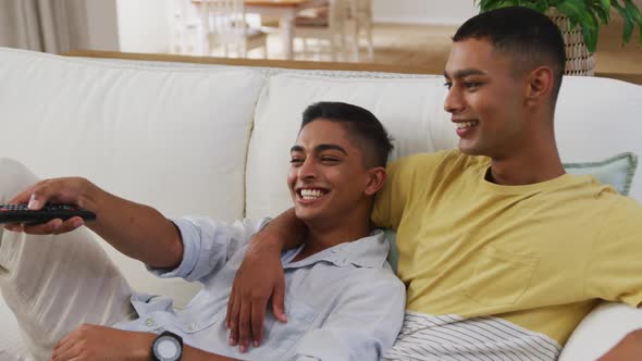 Smiling mixed race gay male couple sitting on sofa watching tv one using remote