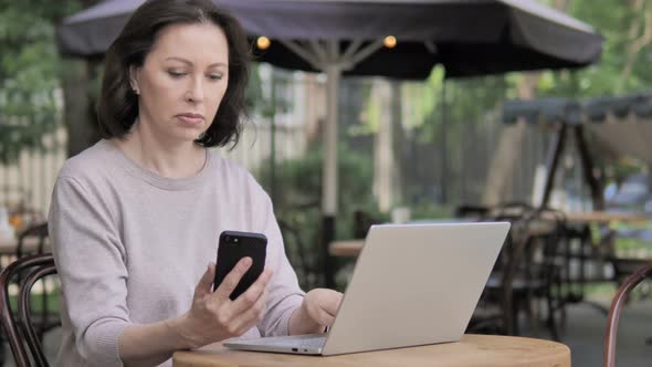 Old Woman Using Smartphone and Laptop, Sitting in Outdoor Cafe