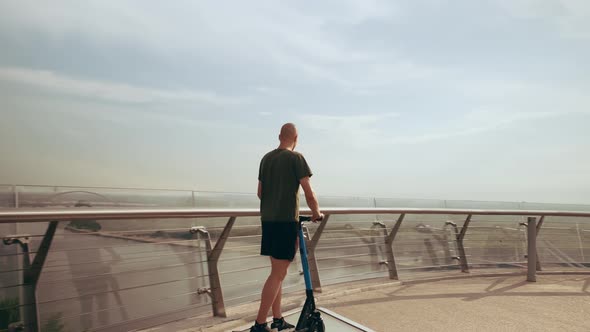 Modern Young Man Rides an Electric Scooter Across the Pedestrian Bridge Overlooking the City