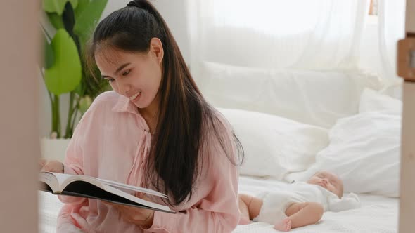 Young Asian mother read book and sit on bed with her newborn baby sleep in background