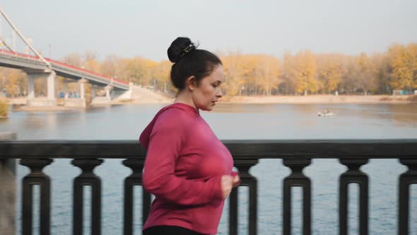 Side follow view of young cute chubby woman easy jogging along city promenade