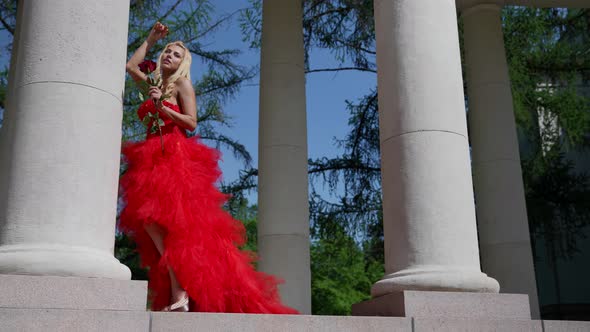 Romantic Shot of Young Blonde Woman in Red Gown with Rose Posing Between White Columns