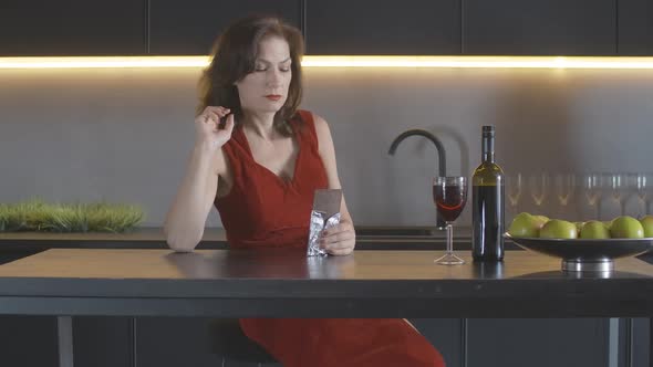 Gorgeous Depressed Mid-adult Woman in Red Dress Sitting in Kitchen and Eating Chocolate. Portrait of