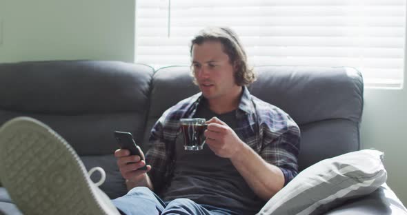 Happy caucasian man sitting on sofa in living room using smartphone and drinking coffee