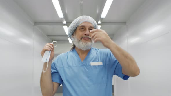 Portrait of Happy Aged Surgeon Removing Safety Mask and Cap Walking in Hospital Corridor