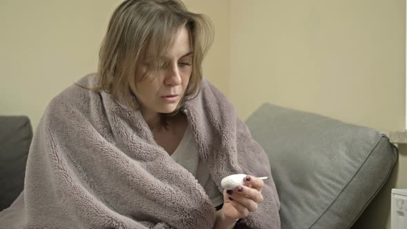 Young Woman Sits Wrapped in a Blanket and Measures the Temperature
