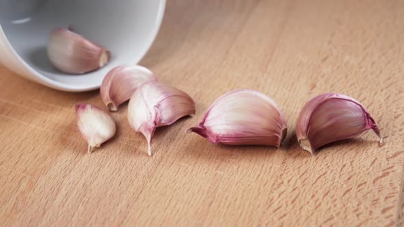 Garlic cloves fall in slow motion from a white cup onto a rustic wooden surface.