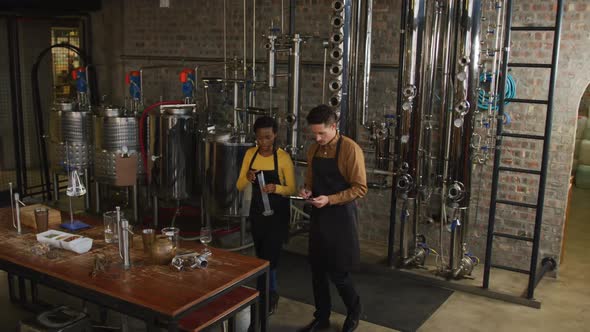 Diverse male and female colleague at gin distillery inspecting equipment, talking and making notes