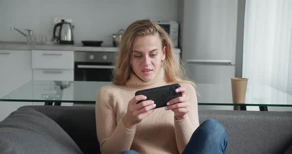 Curly Girl with Blonde Hair Play Game with Smart Phone on Modern Kitchen Background