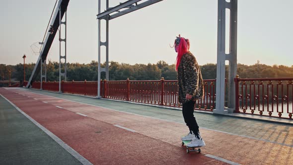 Young Hipster Woman with Pink Hair in Informal Outfit and Protective Mask is Skateboarding on