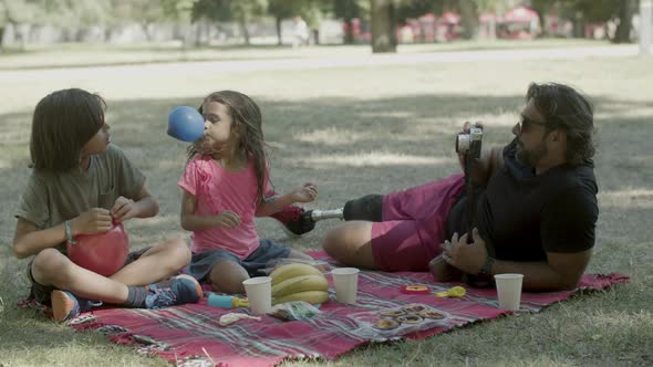 Dad Taking Photo of Kids Inflating Balloons at Picnic