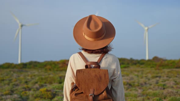 Young traveler woman wearing a hat and with a backpack looking at green alternative turbines