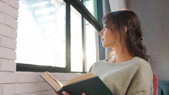 Beautiful Woman Sitting Near Window and Reading a Book on Background of Sunrise